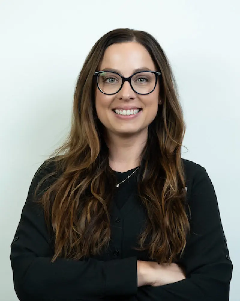 Woman with long brown hair and glasses smiling, wearing a black top, arms crossed.