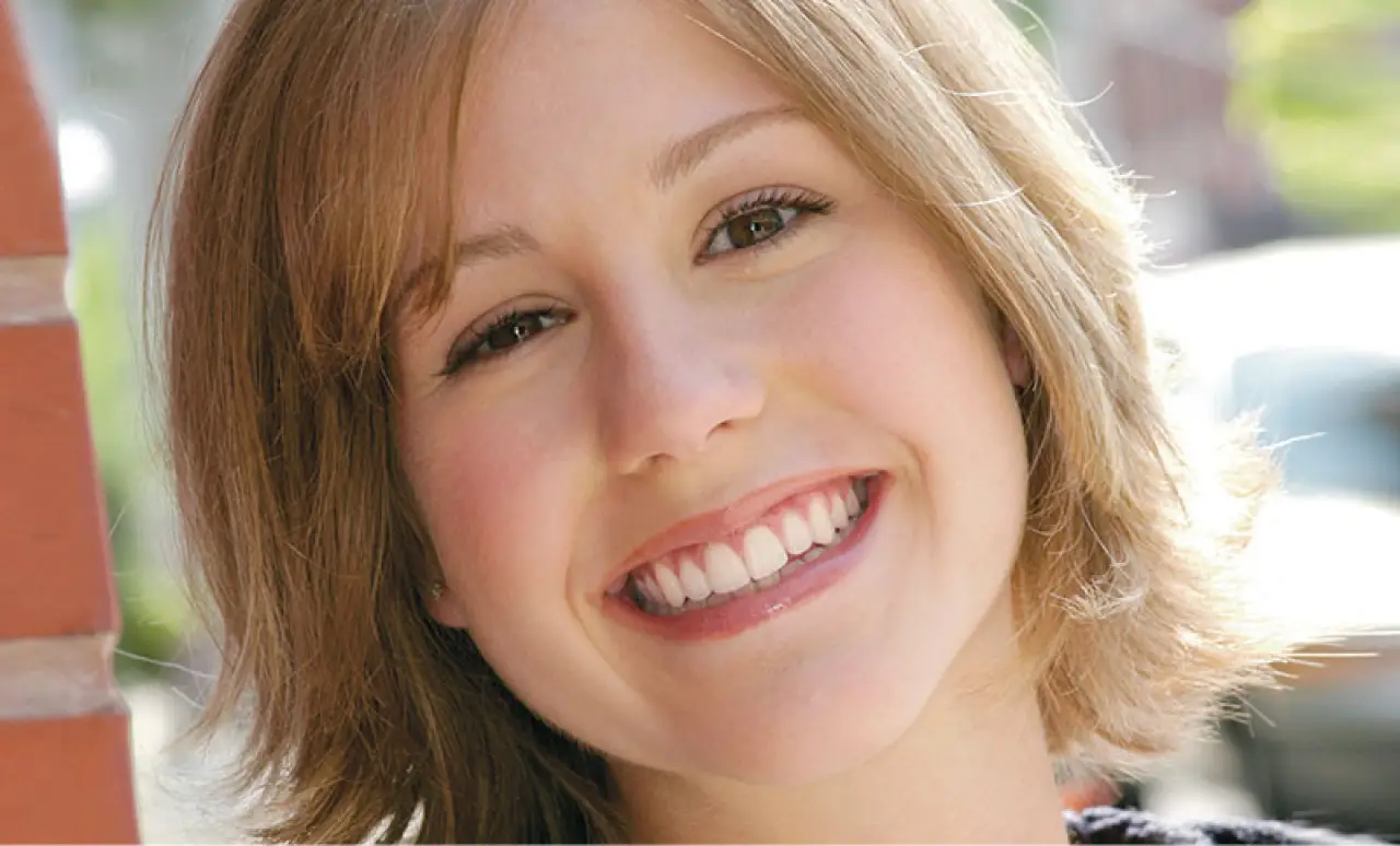 A young woman with short blonde hair smiling outdoors near a brick wall.