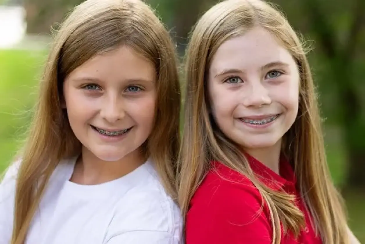 Two smiling girls with long hair and braces pose together outdoors.