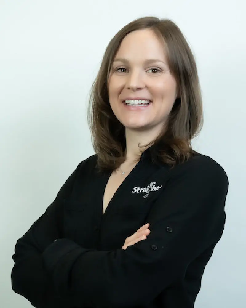 Smiling woman with brown hair in a black shirt, arms crossed, standing against a white background.