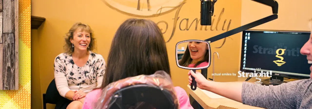 A dentist shows a girl her smile in a mirror while another woman smiles nearby in an office.