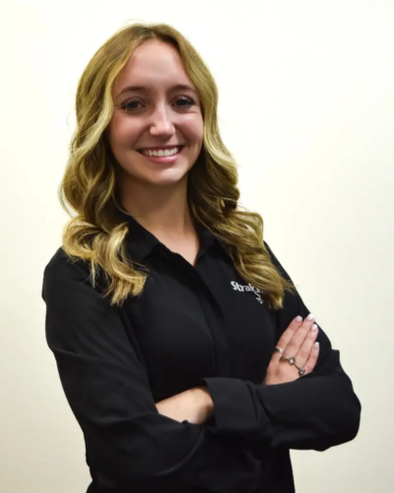Woman with wavy blonde hair in a black shirt, smiling with arms crossed against a white background.