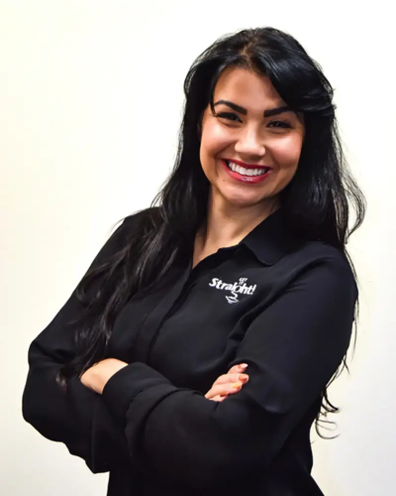 Woman with long dark hair smiling, wearing a black shirt with Straight logo, arms crossed.