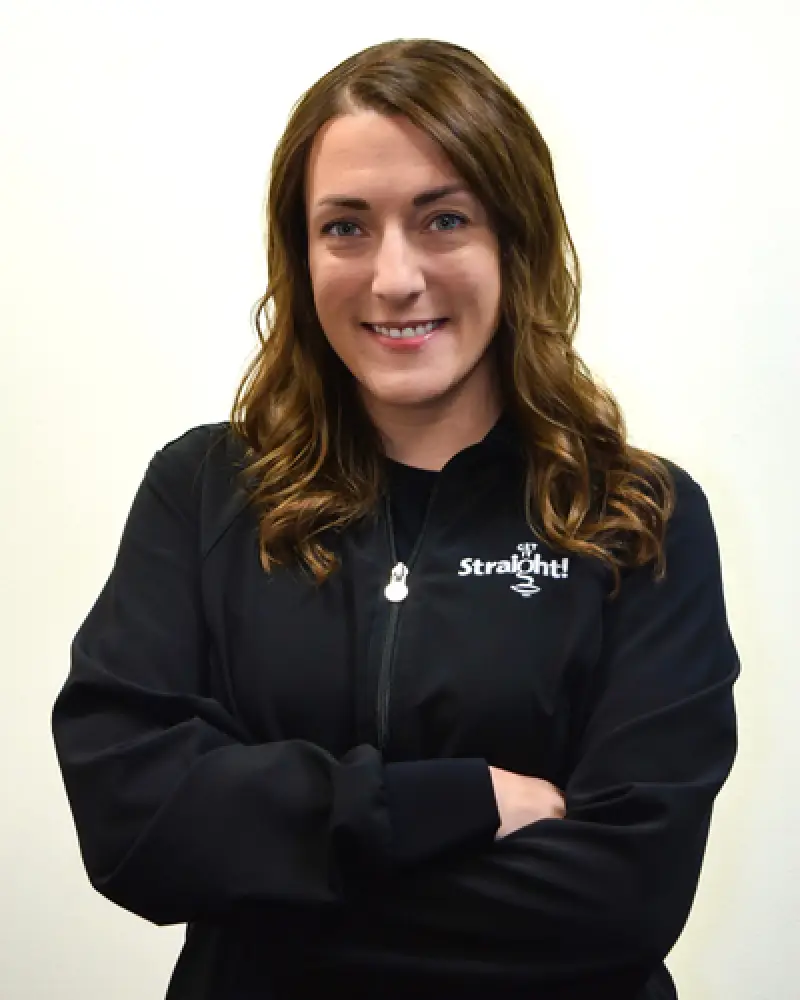 Woman with wavy brown hair in a black Straight jacket, smiling with arms crossed.