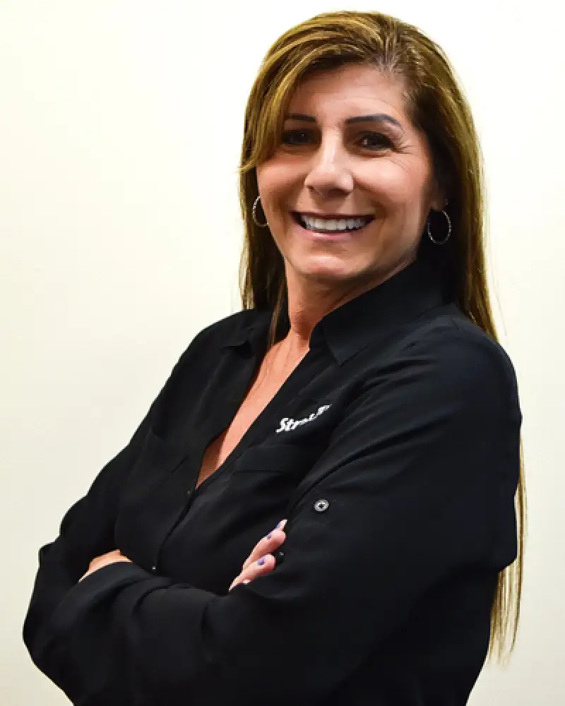 Smiling woman with long brown hair, wearing a black shirt, stands with arms crossed.