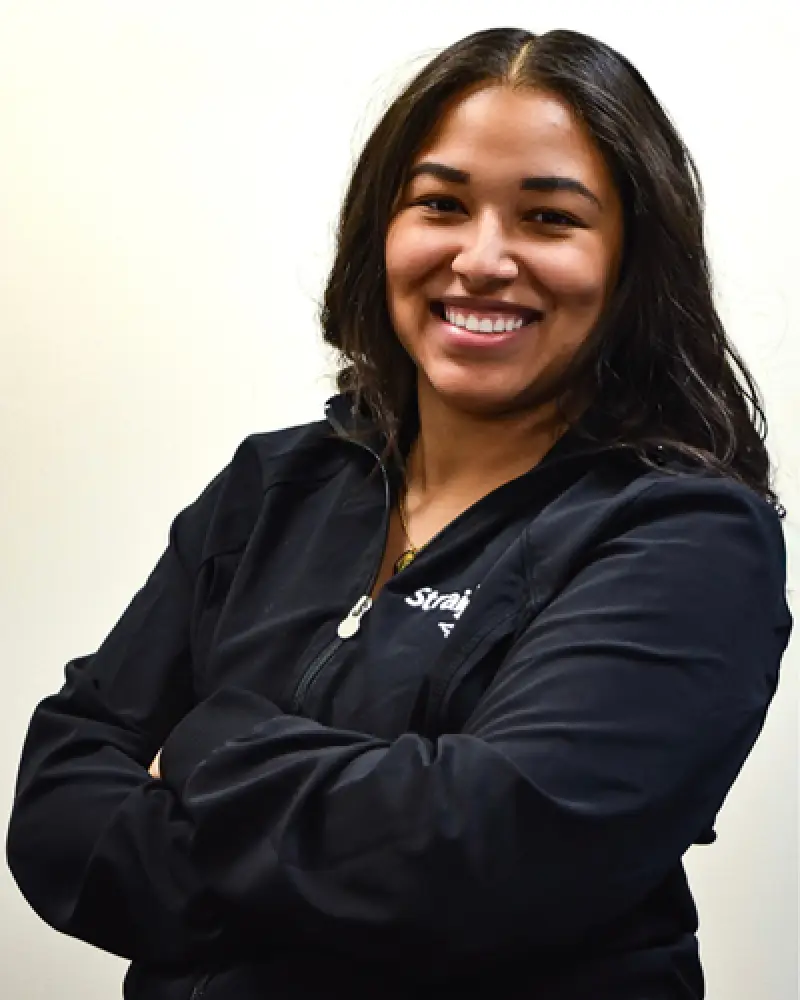 Woman with long dark hair in a black jacket smiling with arms crossed against a light background.