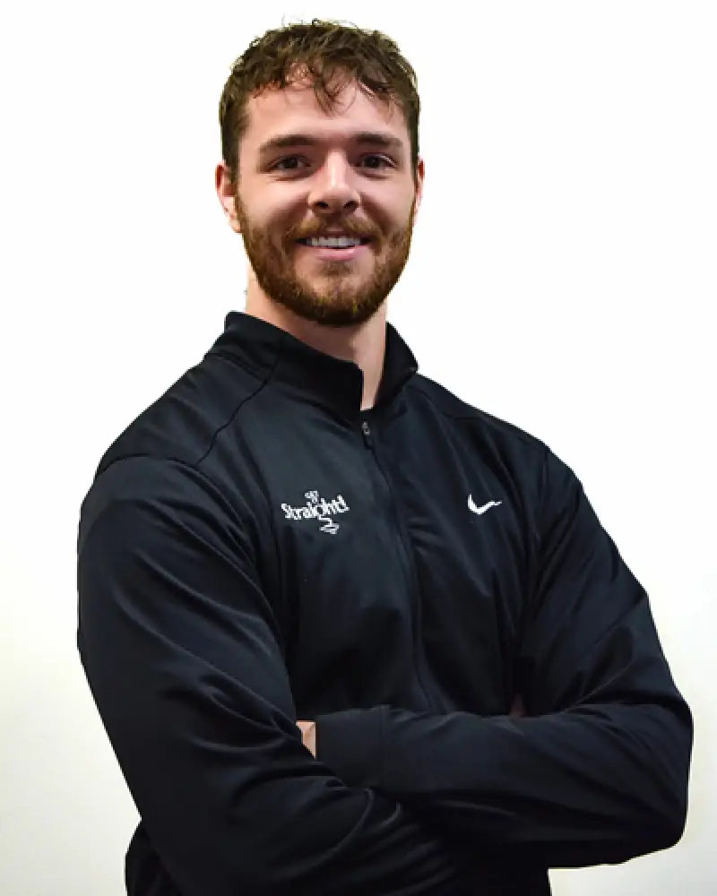 Man with brown hair and beard smiling, wearing a black jacket with arms crossed.