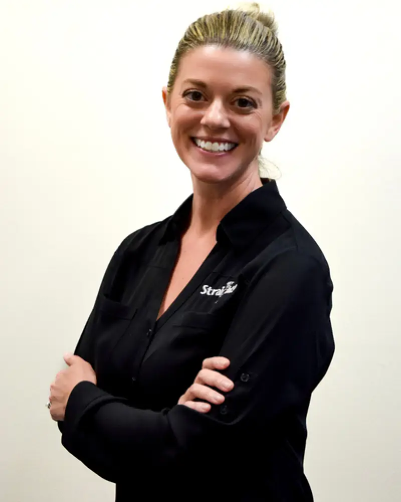 Smiling woman with blonde hair in a black shirt, arms crossed, standing against a white background.