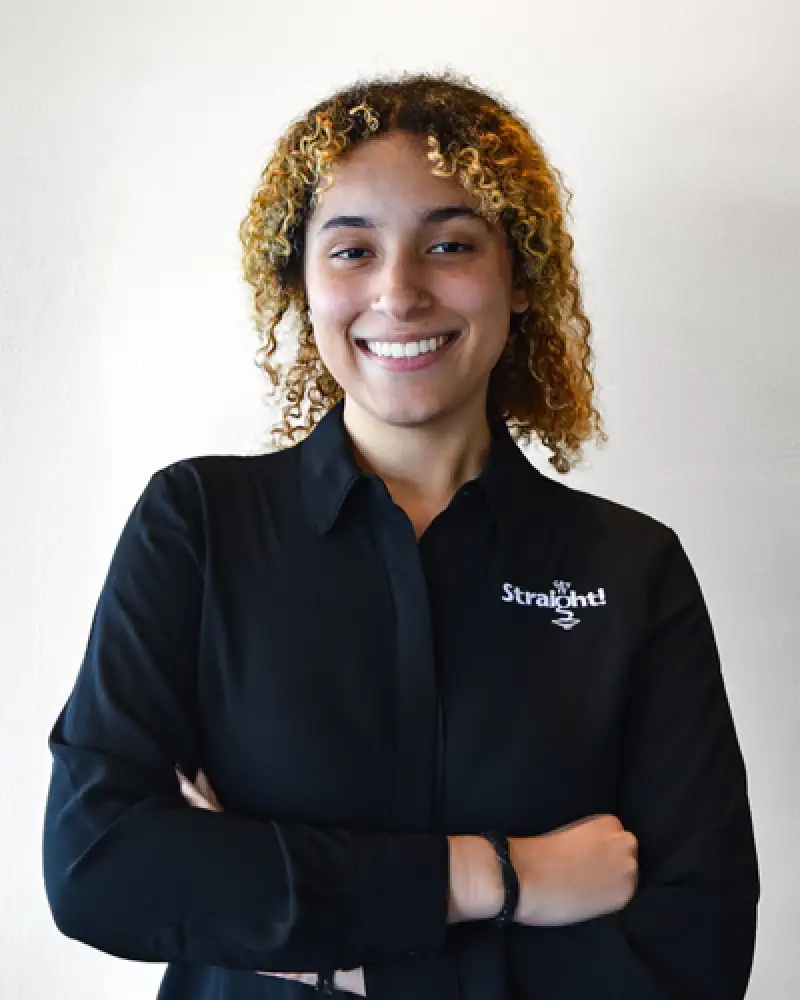 Smiling person with curly hair in a black shirt, arms crossed, standing against a white background.