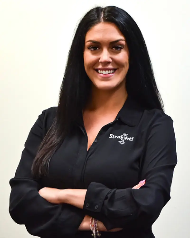 Woman with long dark hair in a black shirt, smiling with arms crossed against a plain background.