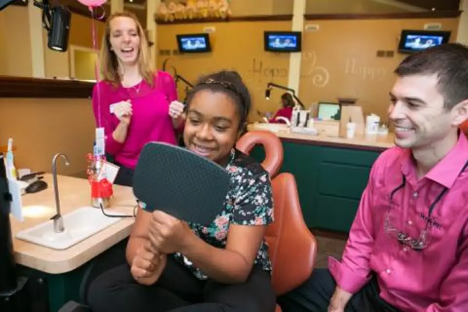 A girl smiles in a dental chair, holding a mirror, with two adults smiling beside her.