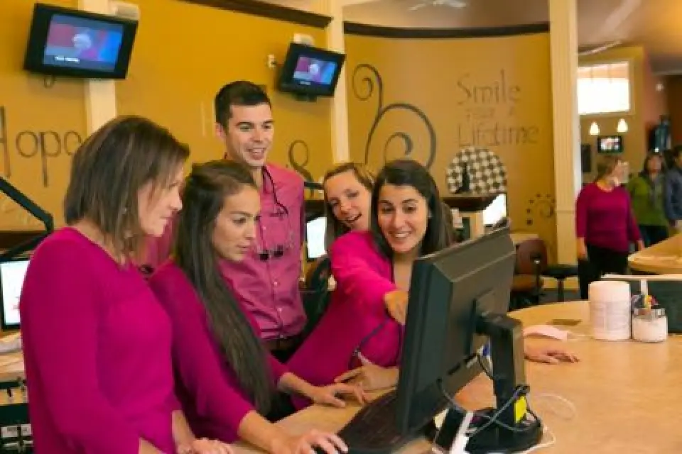 Five people in pink tops smile and gather around a computer at a reception desk in an office.