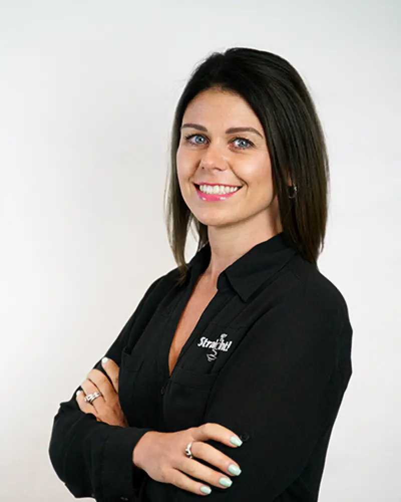 Woman with long brown hair in a black shirt smiling with arms crossed against a white background.