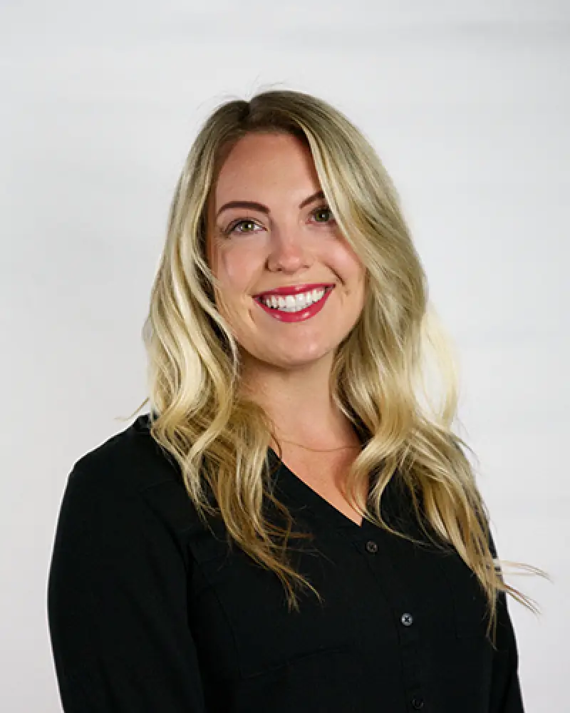 Smiling woman with long blonde hair wearing a black blouse, posed against a plain light background.