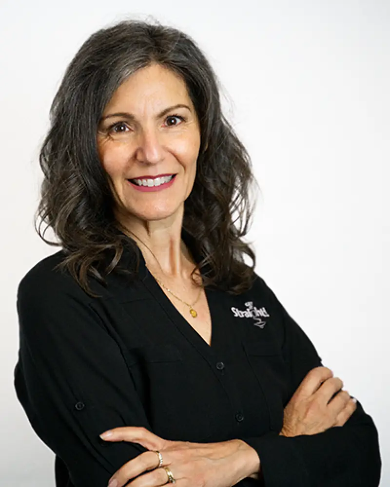 Woman with wavy gray hair, wearing a black shirt, smiling with arms crossed against white background.