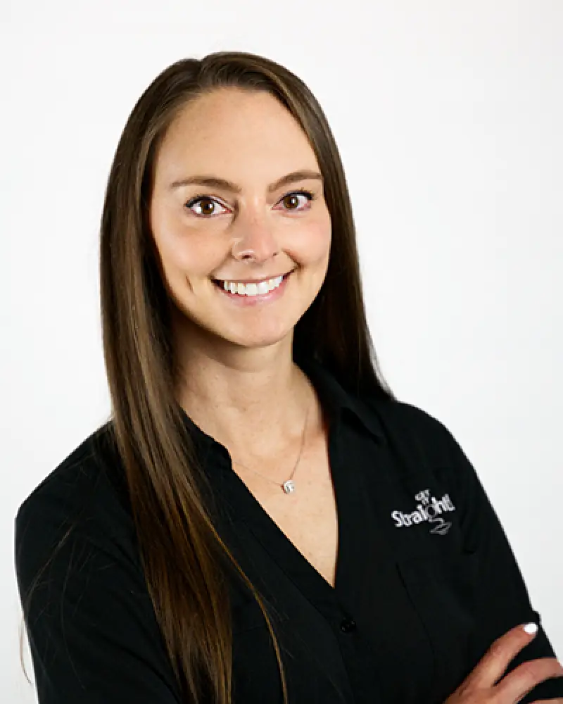 Woman with long brown hair, wearing a black shirt, smiling with arms crossed against white background.