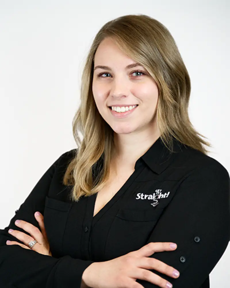 Smiling woman with blond hair in a black shirt stands with arms crossed against a white background.