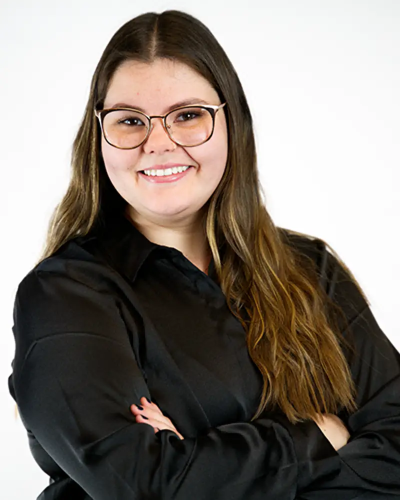 Woman with long brown hair, glasses, and black shirt smiling with arms crossed against white background.