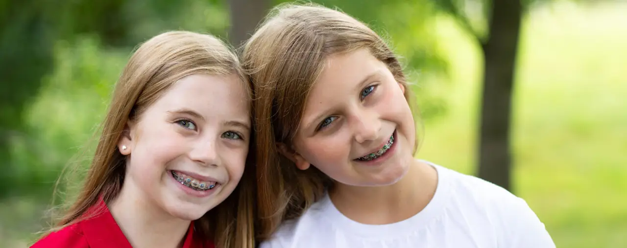 Two girls with braces smiling outdoors, standing close together with trees in the background.