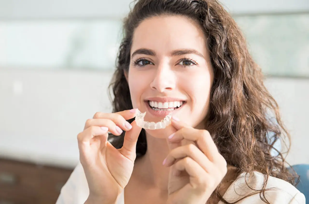 Smiling woman holding a clear dental aligner near her teeth in a bright room.