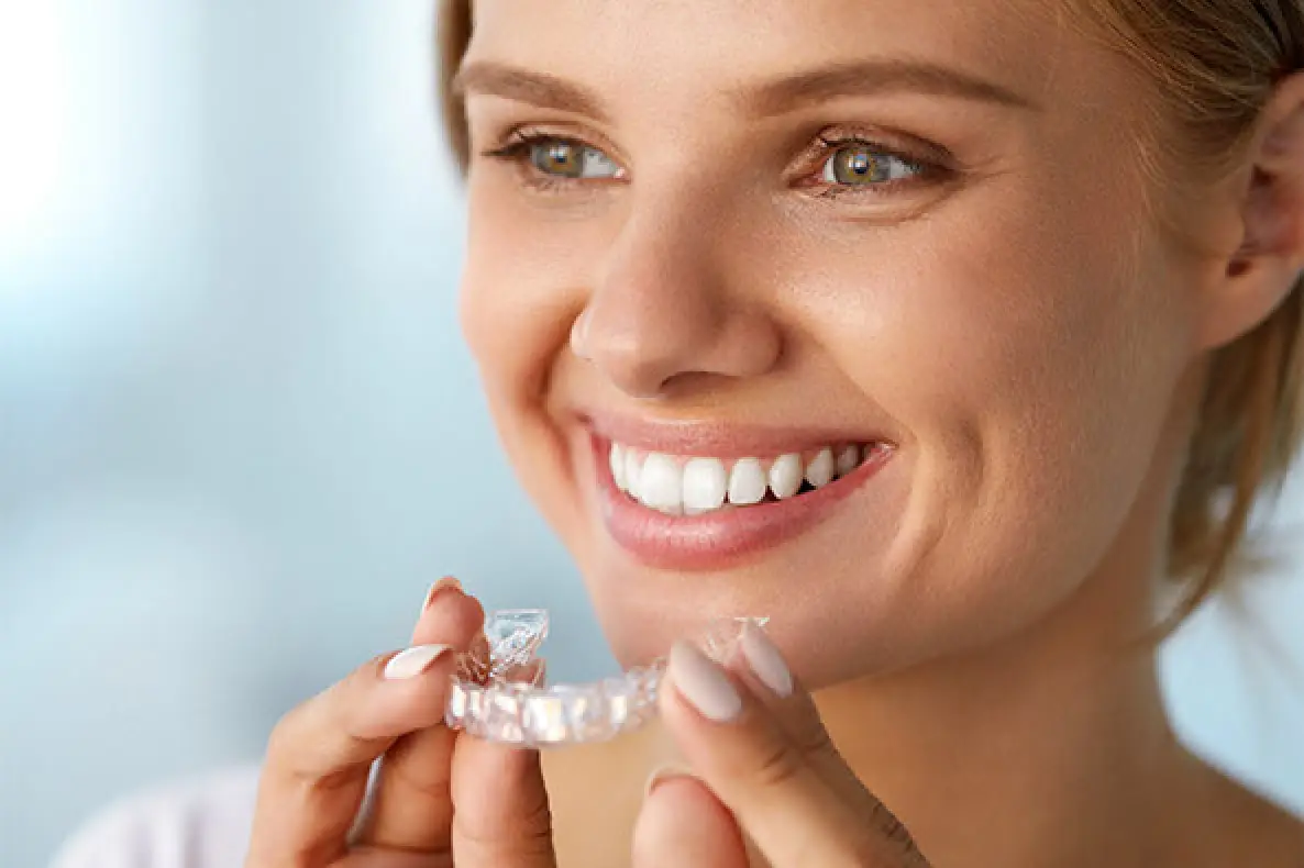 A woman smiles while holding a clear dental aligner, preparing to place it on her teeth.