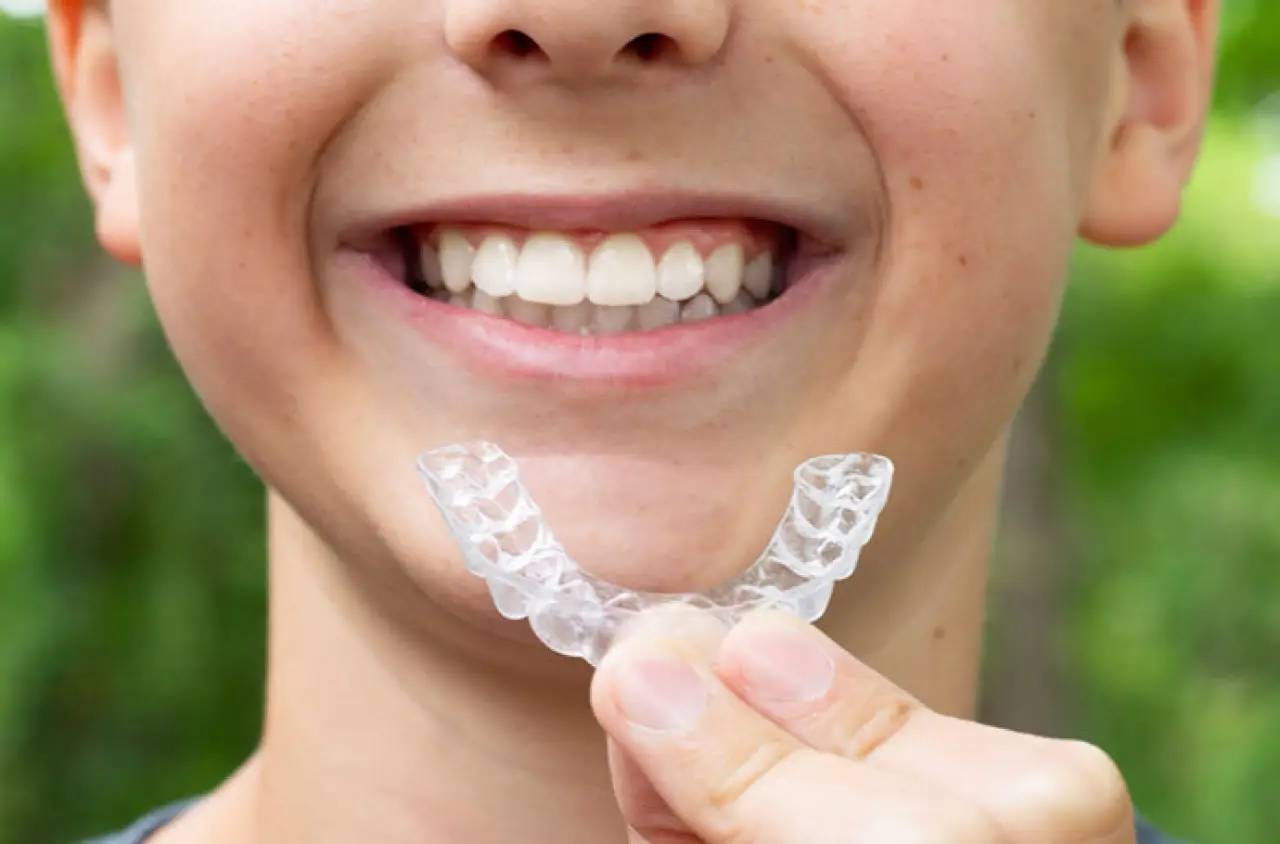 Smiling child holding a clear dental retainer in front of their teeth, outdoors.