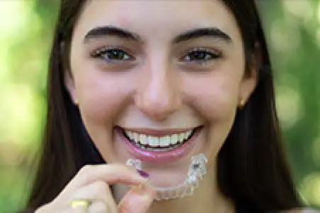 Smiling person holding a clear dental aligner near their teeth, outdoors with greenery behind.