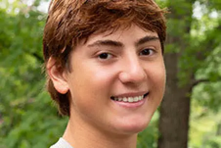 Smiling teen with short brown hair outdoors, green trees and foliage in the background.