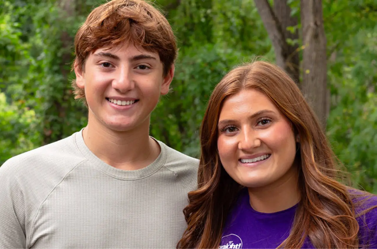 Two smiling young adults standing outdoors in front of green trees.