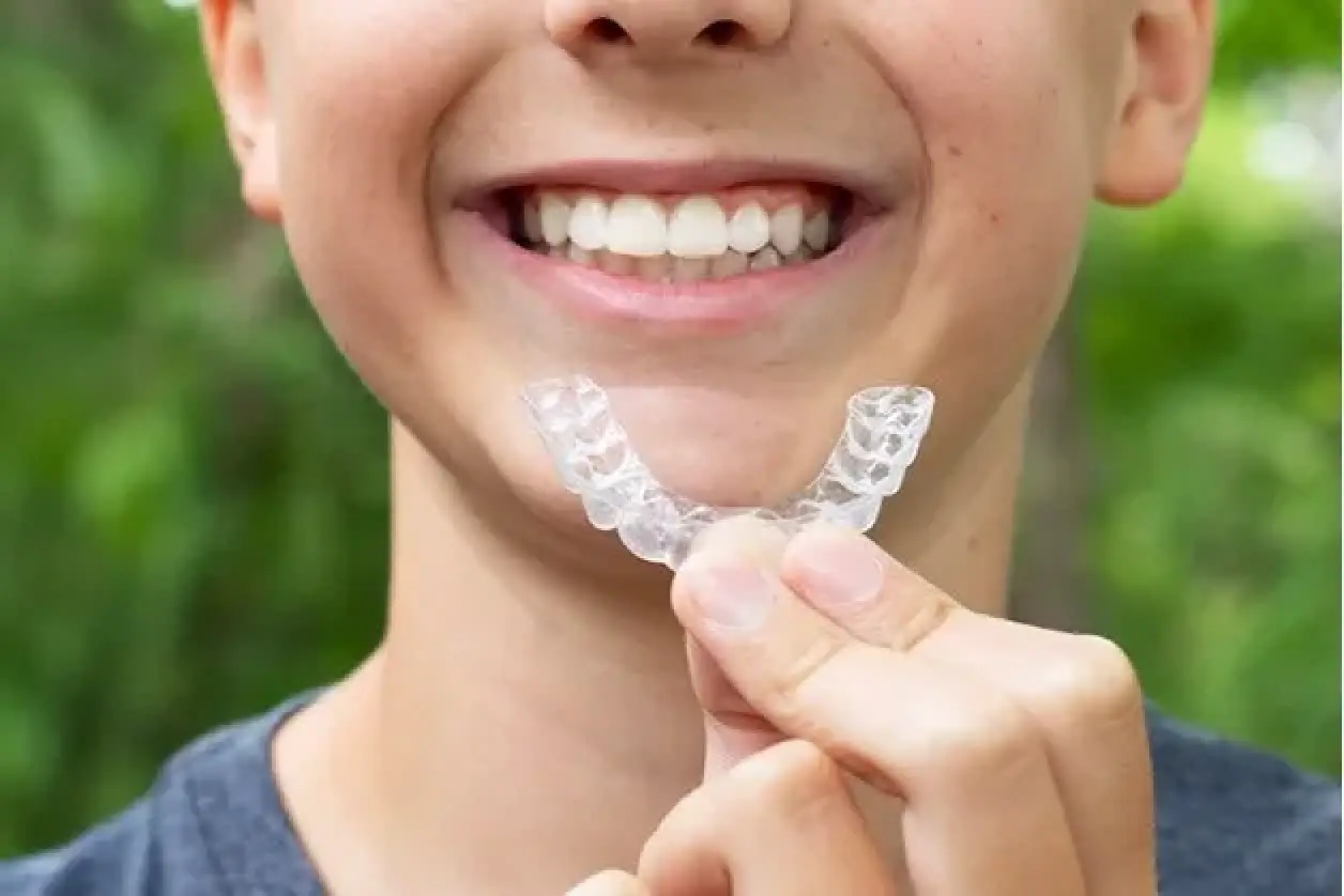 A smiling child holds a clear dental aligner in front of their teeth outdoors.