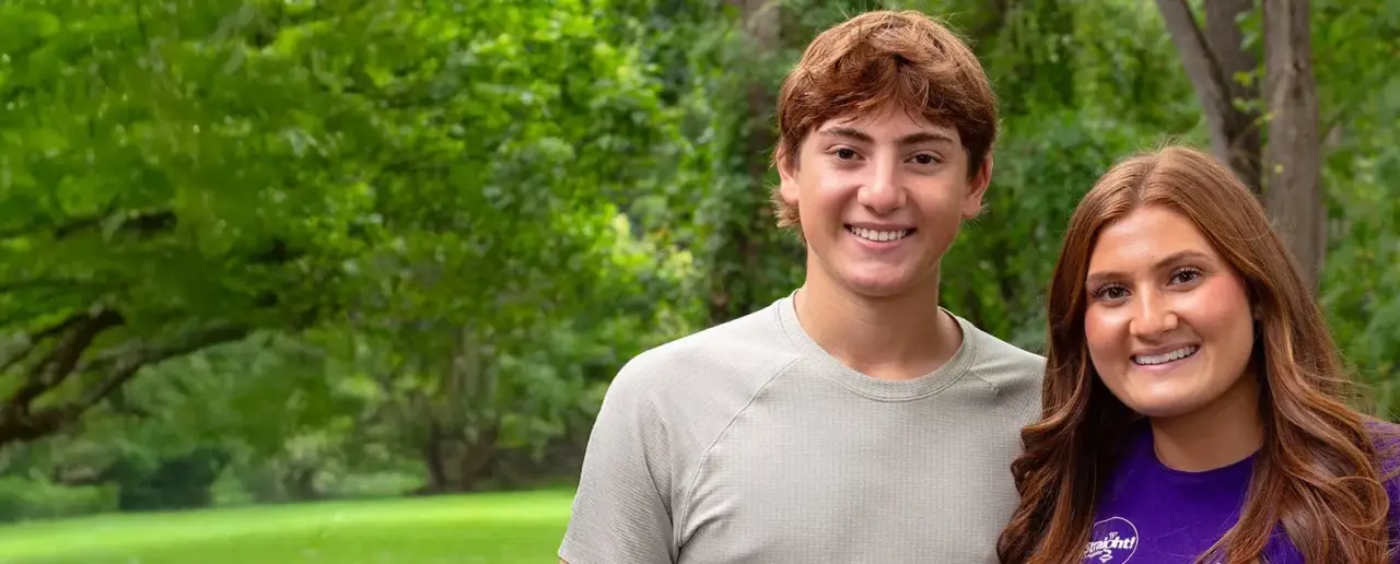 Two smiling young people stand outdoors with trees and grass in the background.