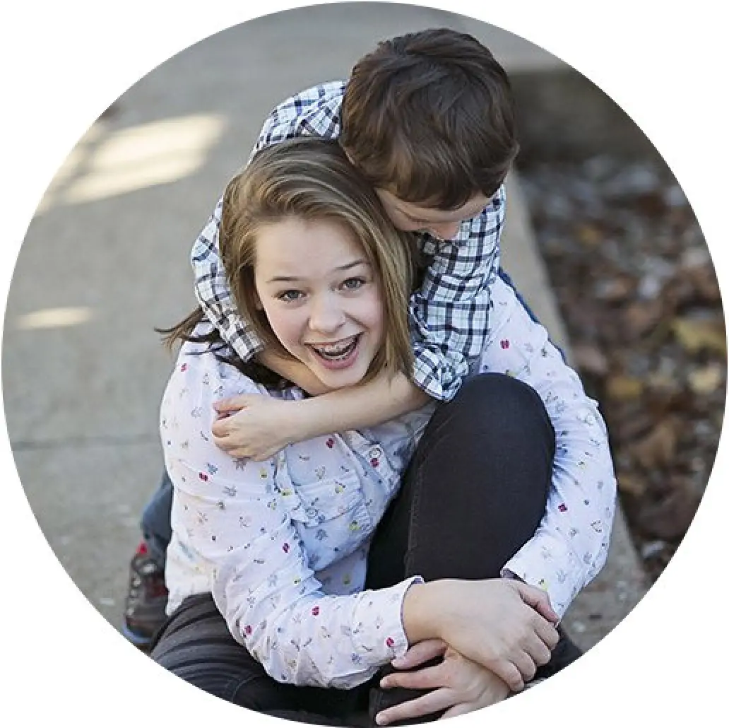Smiling girl sitting as young boy in plaid shirt hugs her from behind outdoors.