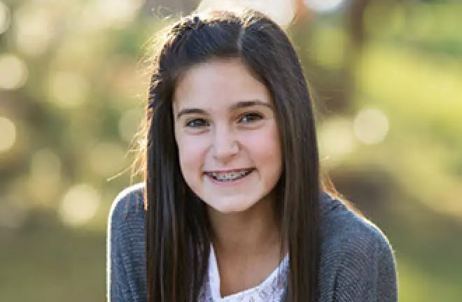 Smiling girl with long dark hair and braces, wearing a gray sweater, outdoors.