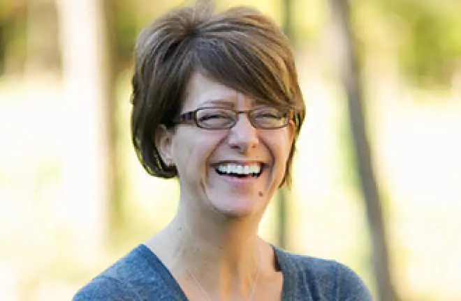 Woman with short brown hair and glasses smiling outdoors with blurred trees in the background.