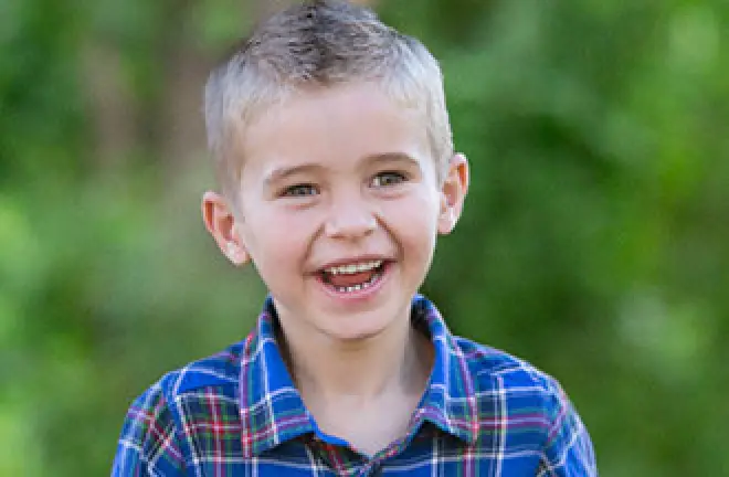 Young boy with short blond hair, smiling and wearing a blue plaid shirt outdoors.
