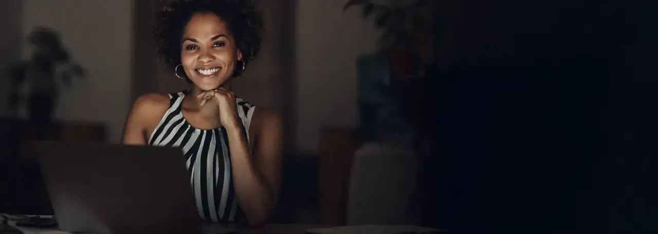 Woman smiling, sitting at a desk with a laptop, resting her chin on her hand.