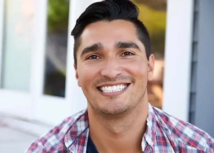 Smiling man with dark hair in a plaid shirt sitting outside in front of windows.
