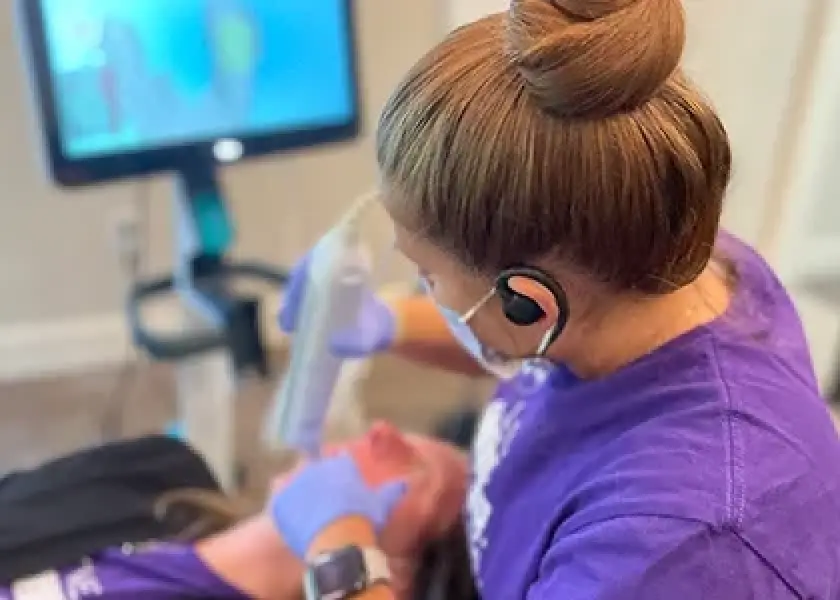 Woman in purple scrubs treats patients face with a device while wearing gloves and a mask.