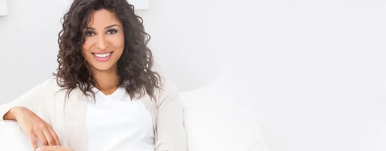 Smiling woman with curly hair sits on a white couch, wearing a white shirt and beige cardigan.