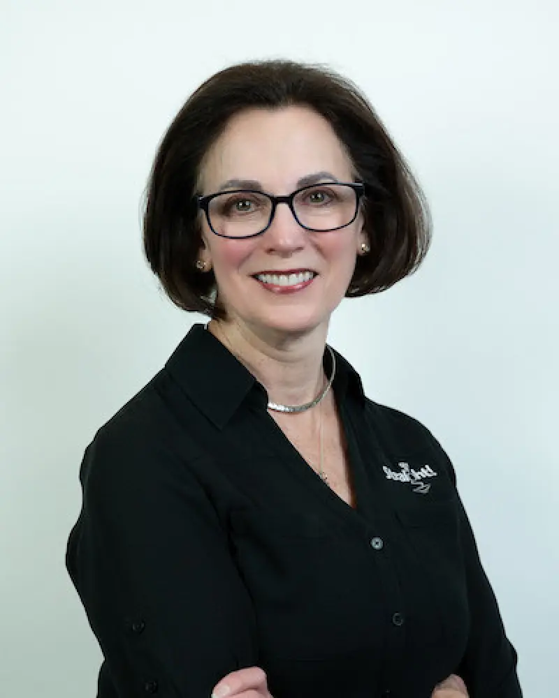 Woman with short dark hair, glasses, and black shirt smiling with arms crossed against white background.