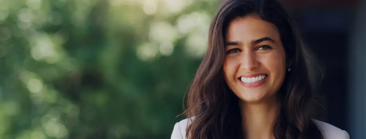 Smiling woman with long dark hair standing outdoors, green foliage blurred in the background.
