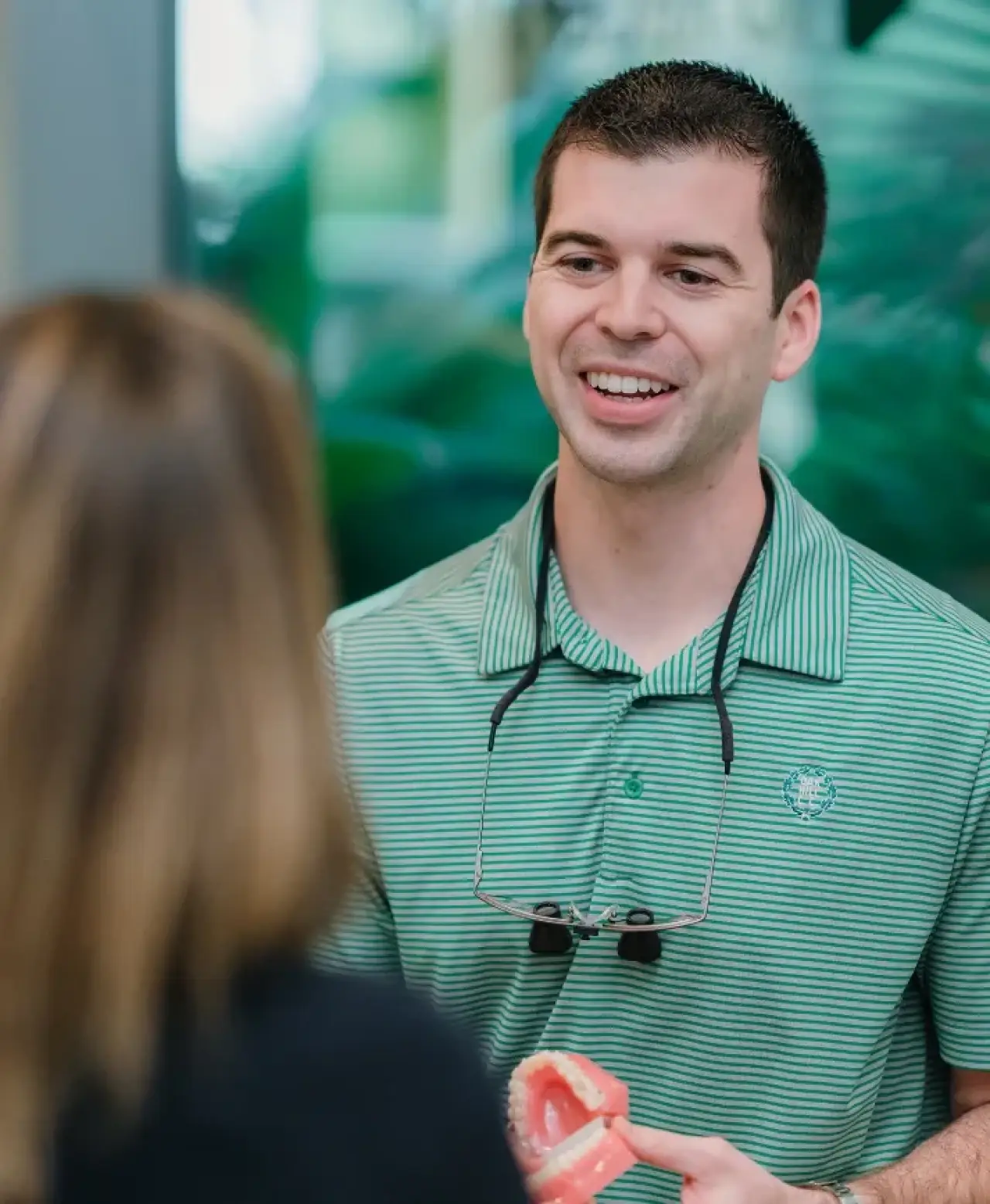 A man in a green shirt smiles while holding dental models and talking to a woman.