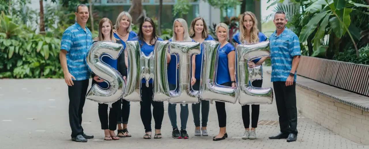 Nine people outdoors holding large silver balloon letters spelling SMILE, all smiling at camera.