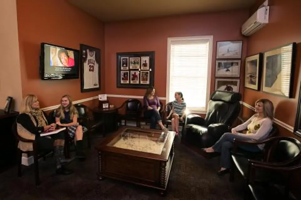 Five women sit and talk in a cozy room with sports memorabilia and a TV on the wall.