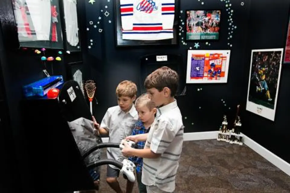 Three young boys play with a joystick exhibit in a sports-themed room with memorabilia on the walls.