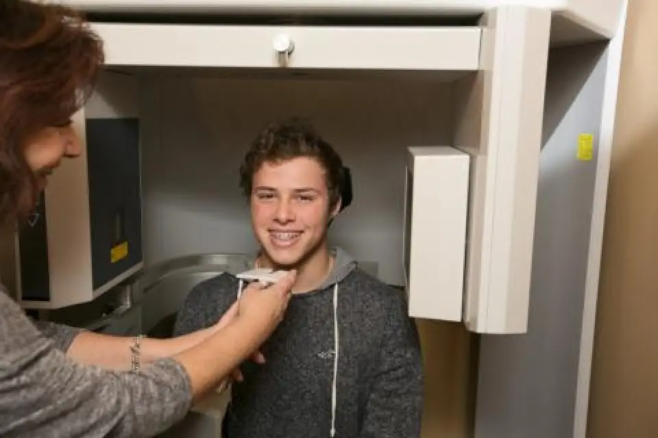 Teenager with braces sits in a dental imaging machine while a woman positions his head.