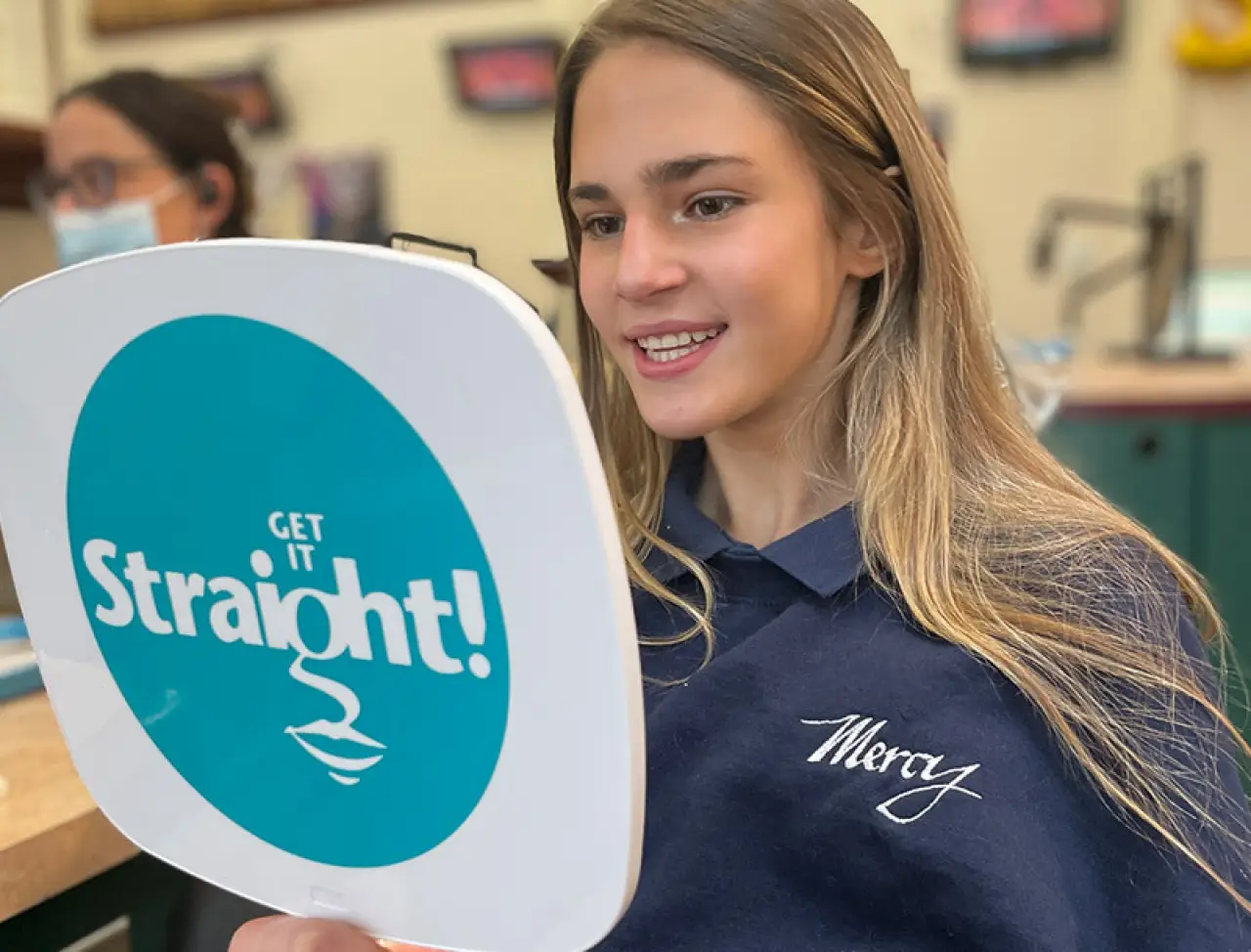 A smiling girl in a Mercy shirt looks into a hand mirror that says Get it Straight