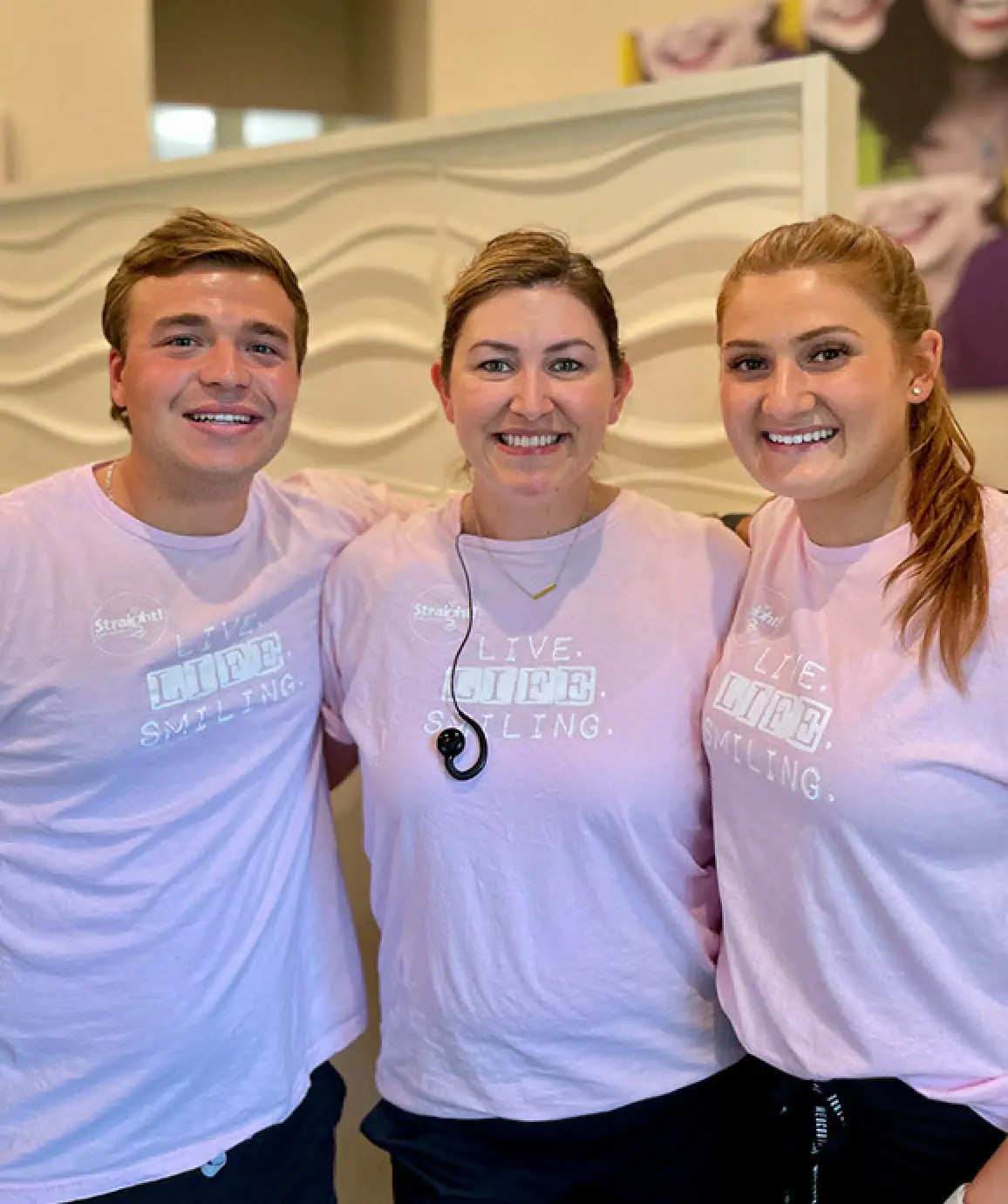 Three people in matching pink Live Life Smiling shirts, smiling and posing together indoors.