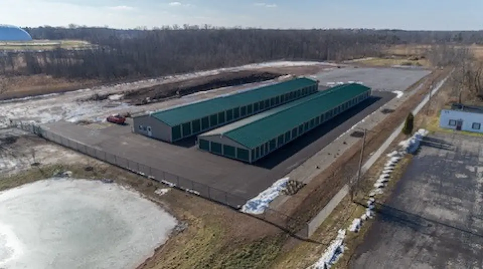 Aerial view of two long green-roofed storage buildings in a fenced lot by a rural road.