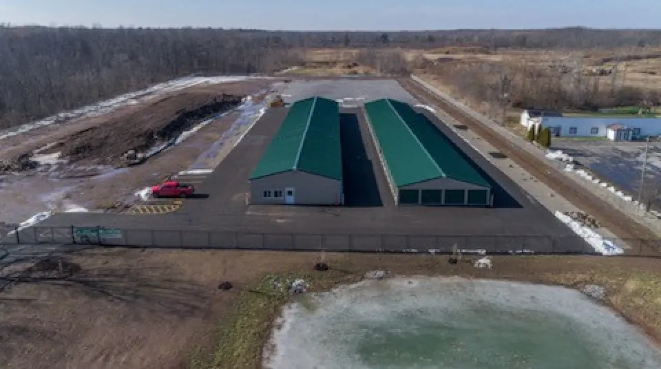 Aerial view of green-roofed storage buildings, a fenced lot, and a pond in a rural area.